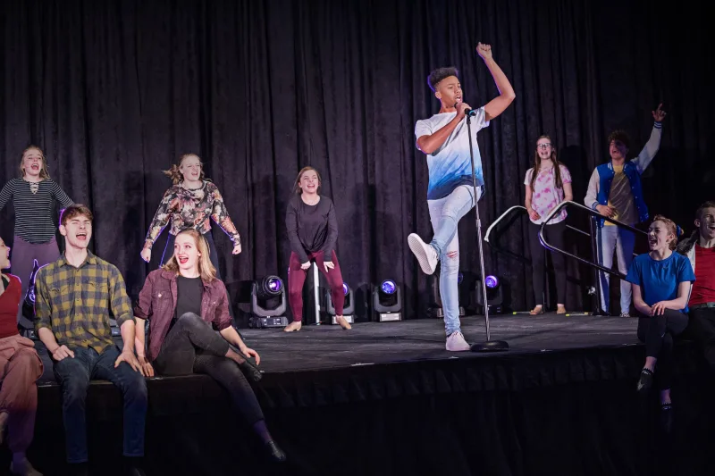 Several actors sitting and standing onstage while singing, with one actor grabbing a microphone on a stand
