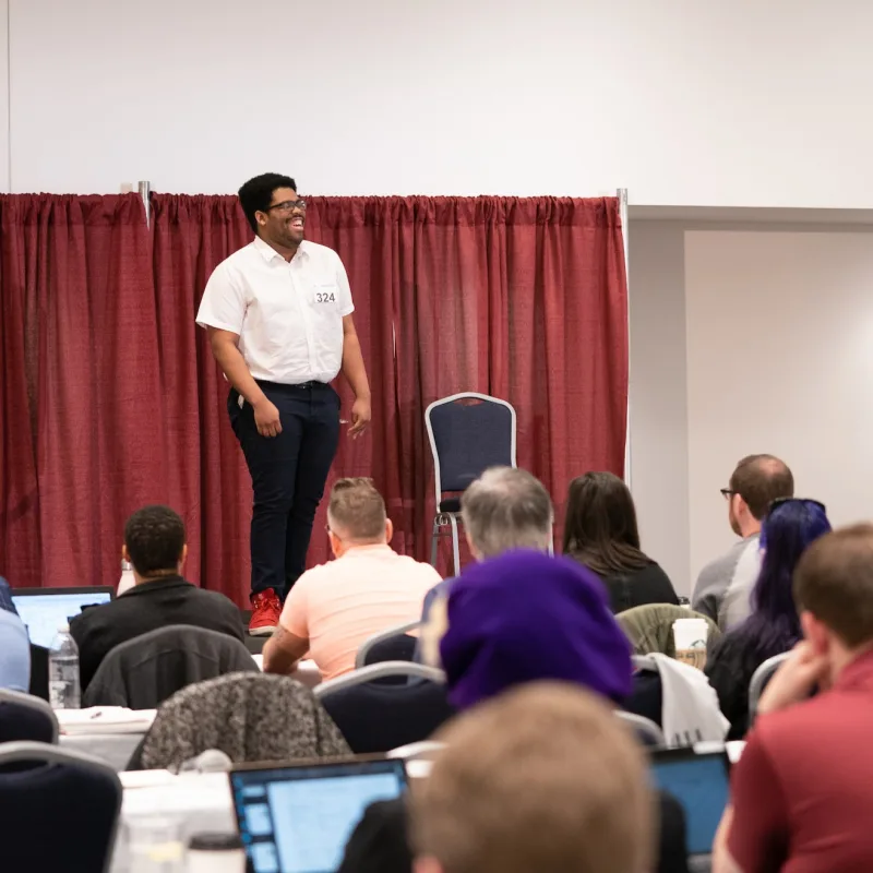 An actor smiling and laughing while standing on a stage in front of auditioners who are seated at tables.